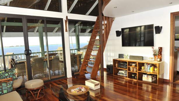 A spacious living room with a large glass door leading to a balcony, a wooden staircase, and a TV mounted on the wall.
