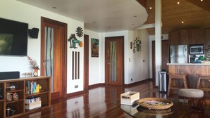 A spacious living room with a wooden floor, a glass coffee table, and a TV on the left.