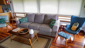 A living room with a grey sofa, a blue armchair with a pineapple pillow, a wooden coffee table, and a small round table with a stool.