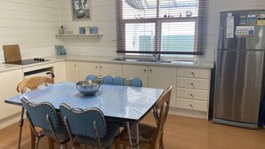 Une cuisine équipée d’une table à manger, de chaises, d’un réfrigérateur, d’une fenêtre avec des stores et d’une étagère contenant des ustensiles de cuisine.