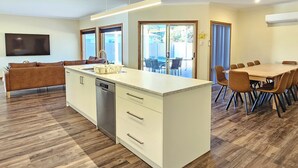 Eine moderne Küche mit einer zentralen Kochinsel, ein Essbereich mit großem Tisch und Stühlen sowie eine Wohnbereits mit Sofa und Fernseher.