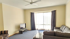 Ein Wohnzimmer mit Flachbildfernseher, zwei Sofas, Deckenventilator und großen Fenstern mit Vorhängen.