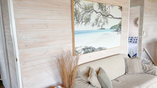 Una habitación con un sofá color beige, un cuadro enmarco con una escena de playa y una mesita de mimbre con revistas.