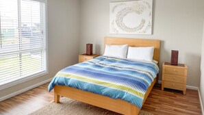 A bedroom with a bed, wooden nightstands, a large window with blinds, and a wall-mounted artwork.