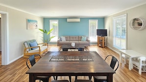 A spacious living room with a large dark wood dining table, a grey sofa, and a TV stand.