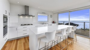 A modern kitchen with white cabinetry, a large island with bar stools, a gas stove, and a view of the sea through large windows. - Bayview - Fantastic Panoramic Views of Guichen Bay - Pet Friendly Inside (Robe)