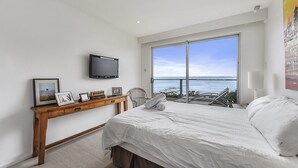 A bedroom with a large bed, a TV mounted on the wall, a wooden console table with framed photos, and a large window offering a view of the sea. - Bayview - Fantastic Panoramic Views of Guichen Bay - Pet Friendly Inside (Robe)