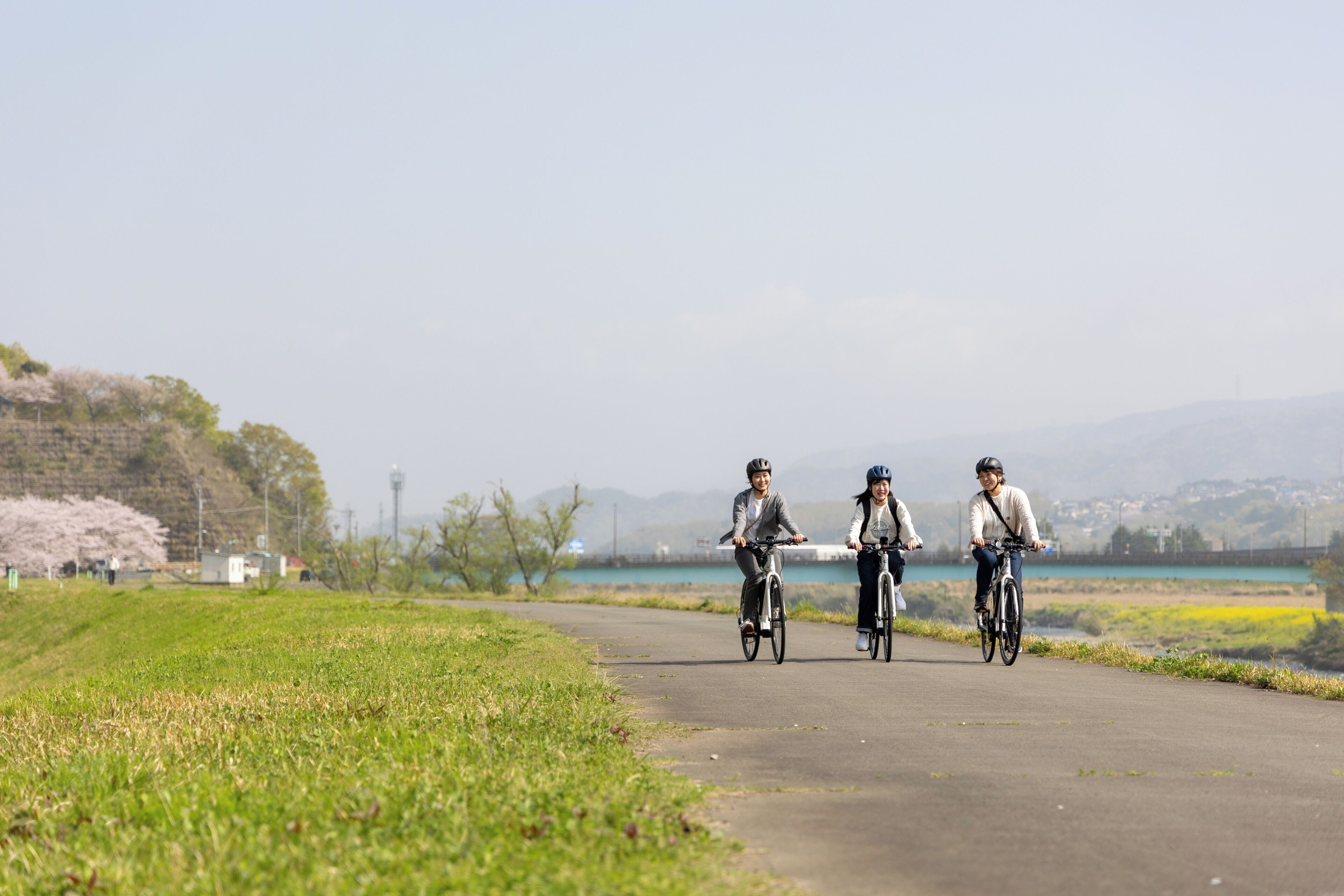 Three people riding bicycles on a paved path with greenery and a body of water in the background.