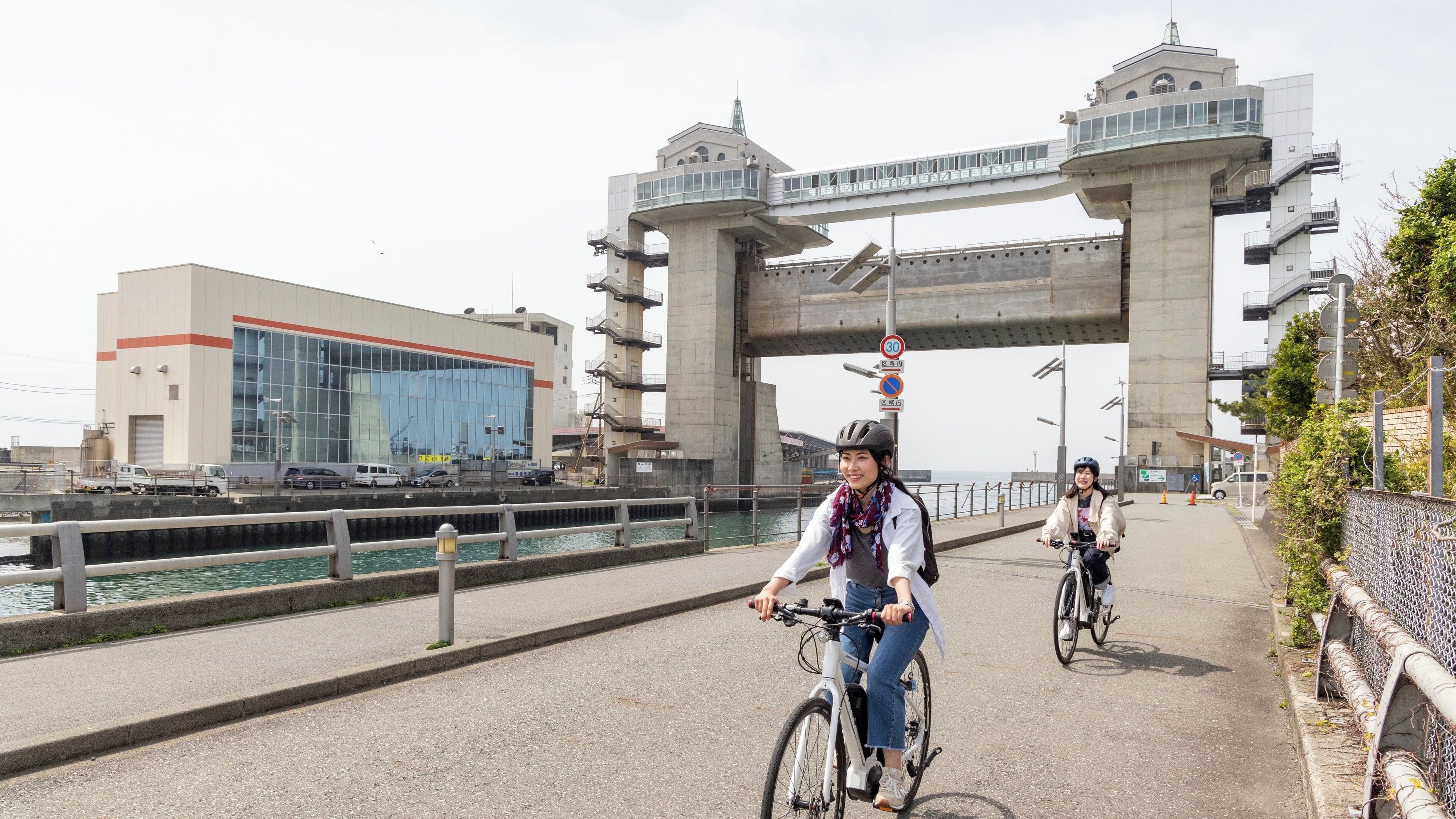Un pont per a vianants sobre un canal amb un edifici modern a l'esquerra i una tanca metàl·lica a la dreta.