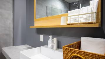 A bathroom with a white sink, a mirror above it, a wooden shelf with toiletries, and a wicker basket with a towel.