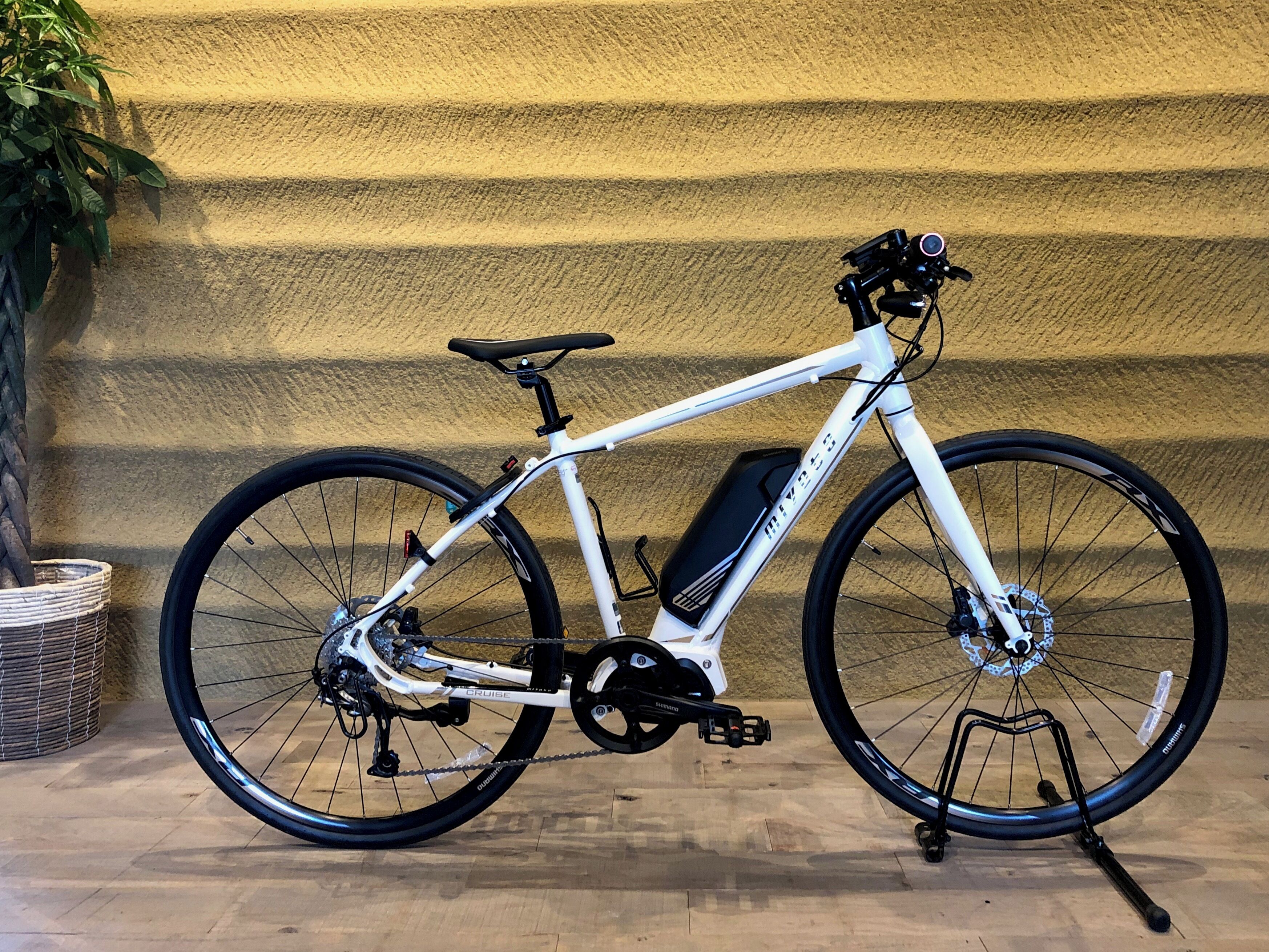 Una bicicleta blanca con asiento negro y canasta delantera, estacionada frente a una pared amarilla con textura.
