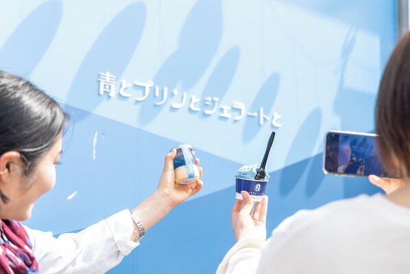 A person holding a cup with a straw, another person taking a photo with a smartphone, and a blue background with Japanese text. - Konastay Izu-Nagaoka - Hostel (Izunokuni)