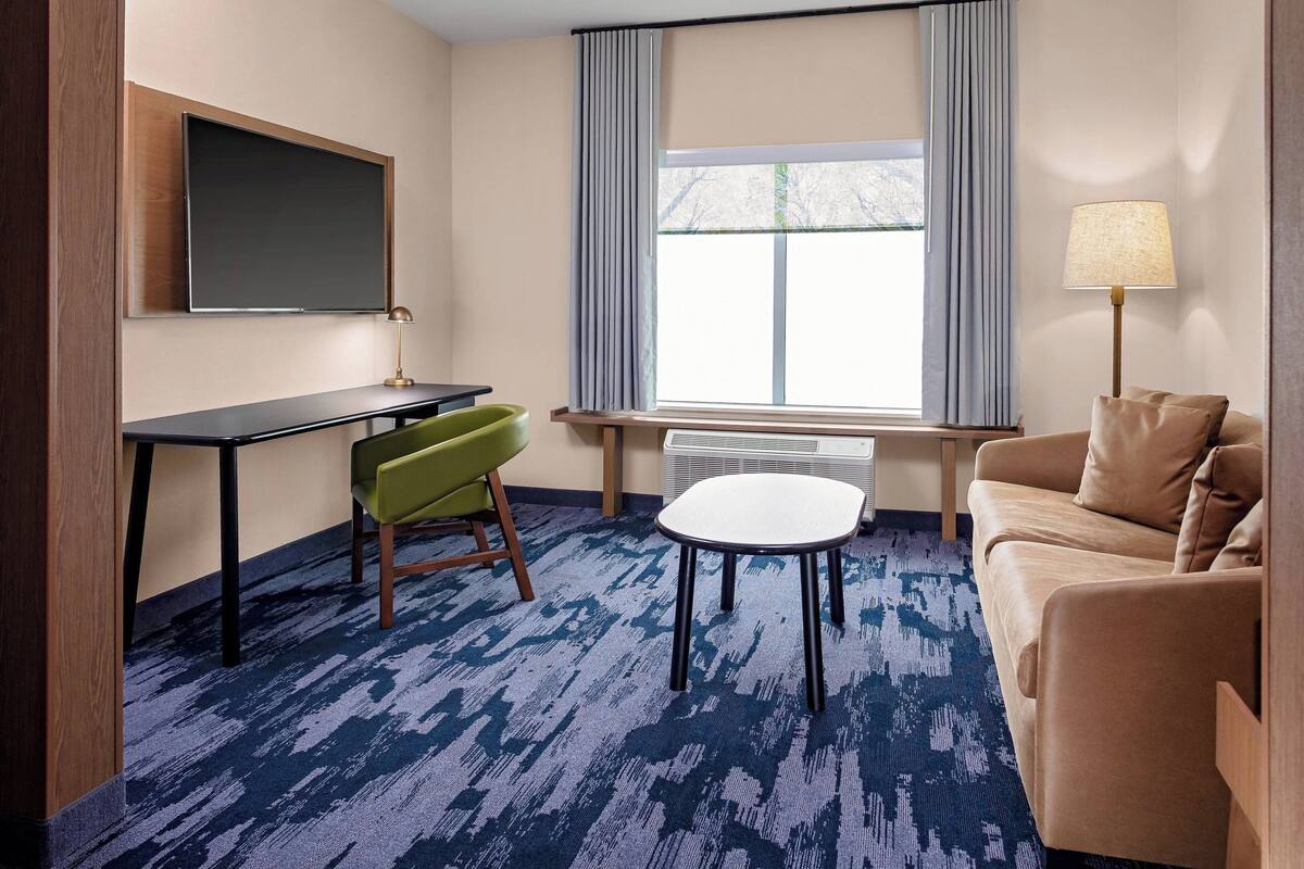 A hotel room with a desk, green chair, white table, beige sofa, and a large window with curtains.