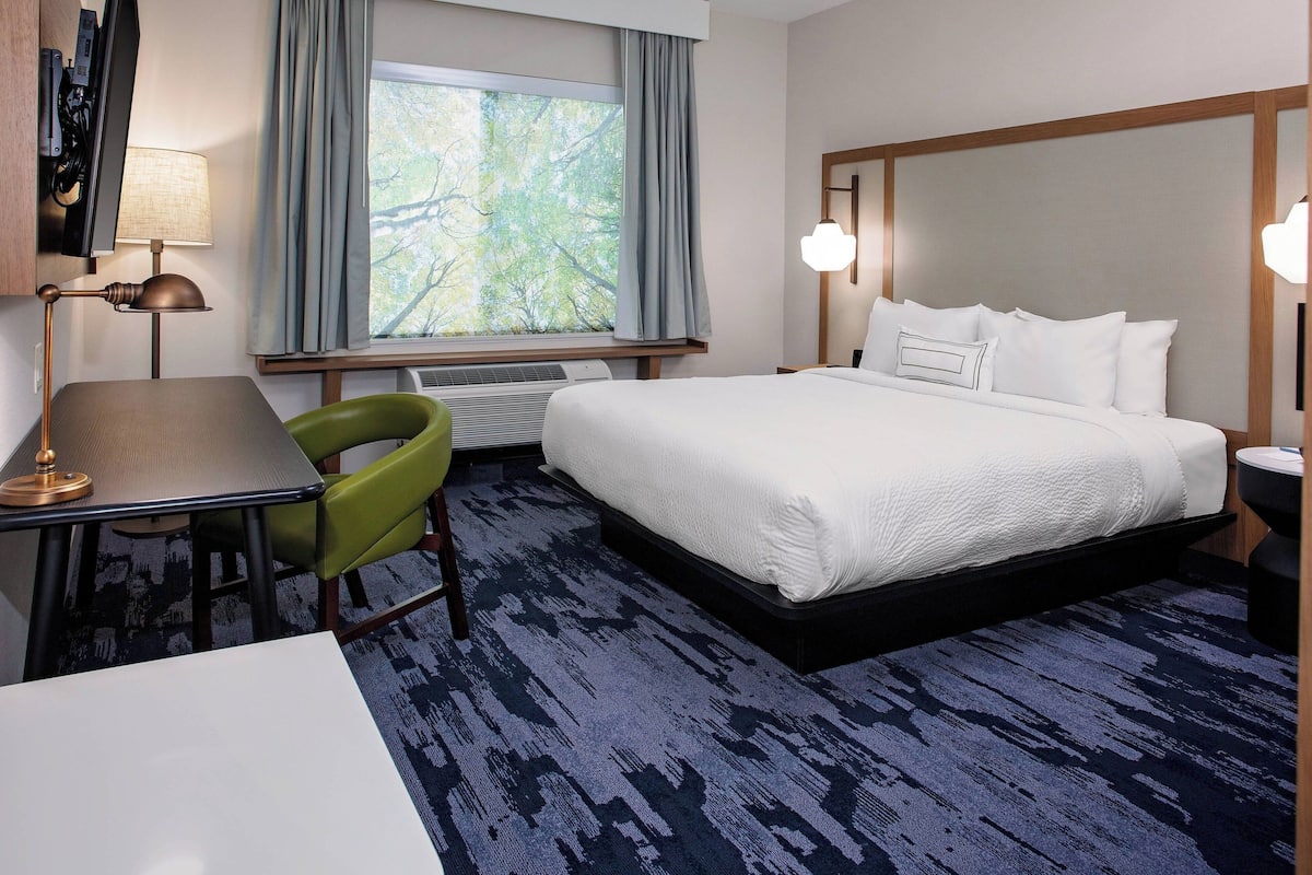 A hotel room with a bed, a desk with a lamp, a chair, a window with curtains, and a nightstand with a lamp.