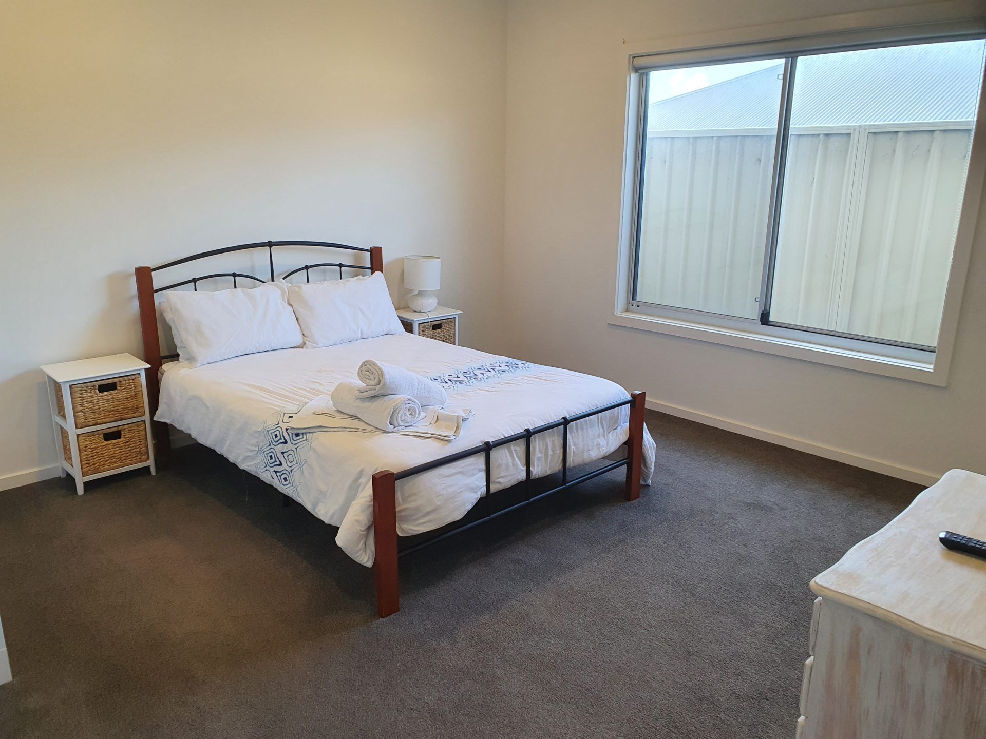 A neatly made bed with white linens and a rolled towel, a bedside table with a lamp, and a window with a view of a white fence.