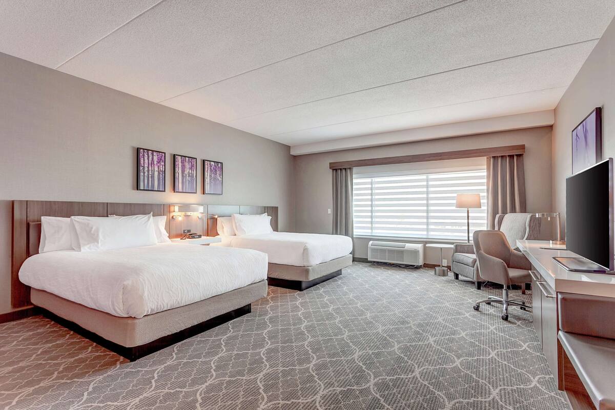 Une chambre d’hôtel avec deux lits, un bureau avec une chaise, un téléviseur à écran plat, une fenêtre avec des stores et des œuvres d’art accrochées au mur.