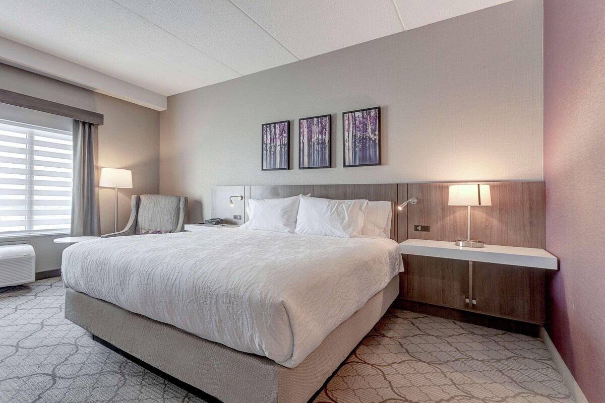A hotel room with a large bed, bedside tables, a desk, and a window with blinds.