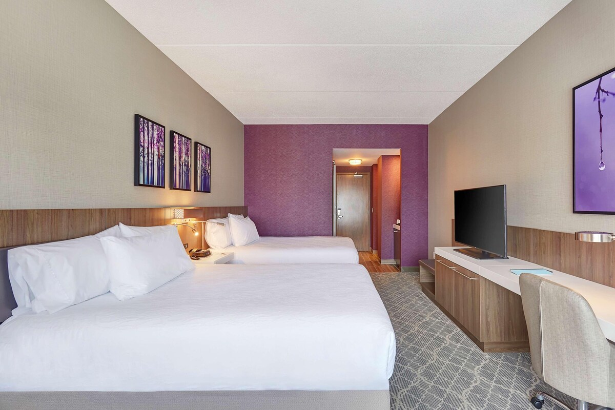 A hotel room with two beds, a desk with a TV, and artwork on the walls.