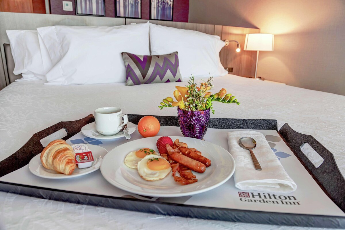 A neatly arranged breakfast tray on a bed with a pillow, a vase with flowers, and a tray with croissants, eggs, bacon, and an orange.