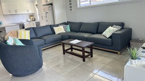 Un salon moderne avec un canapé d’angle, un fauteuil rond bleu, une table basse en bois et une cuisine en arrière-plan.