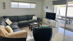 Un salon avec un canapé gris, une table basse, un téléviseur et une porte-fenêtre coulissante donnant sur une terrasse extérieure.