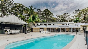 Piscina com espreguiçadeiras, área de estar coberta e um edifício com grandes janelas e telhado metálico.