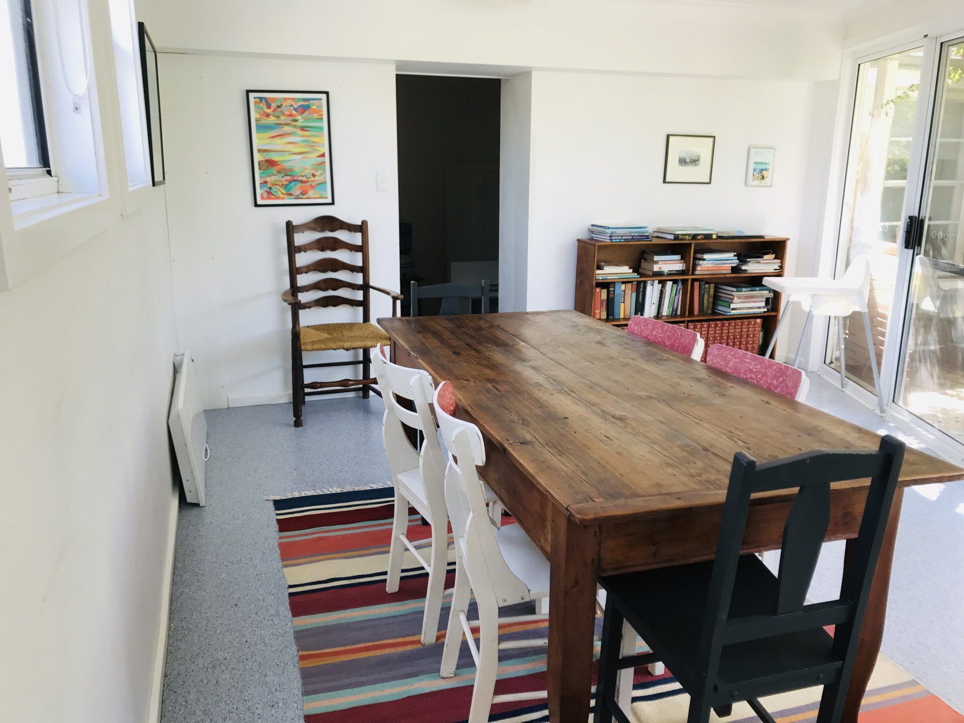 Ein Essbereich mit einem Holztisch, weißen Stühlen, einem farbenfrohen Streifen Teppich und einem Bücherregal mit Büchern.