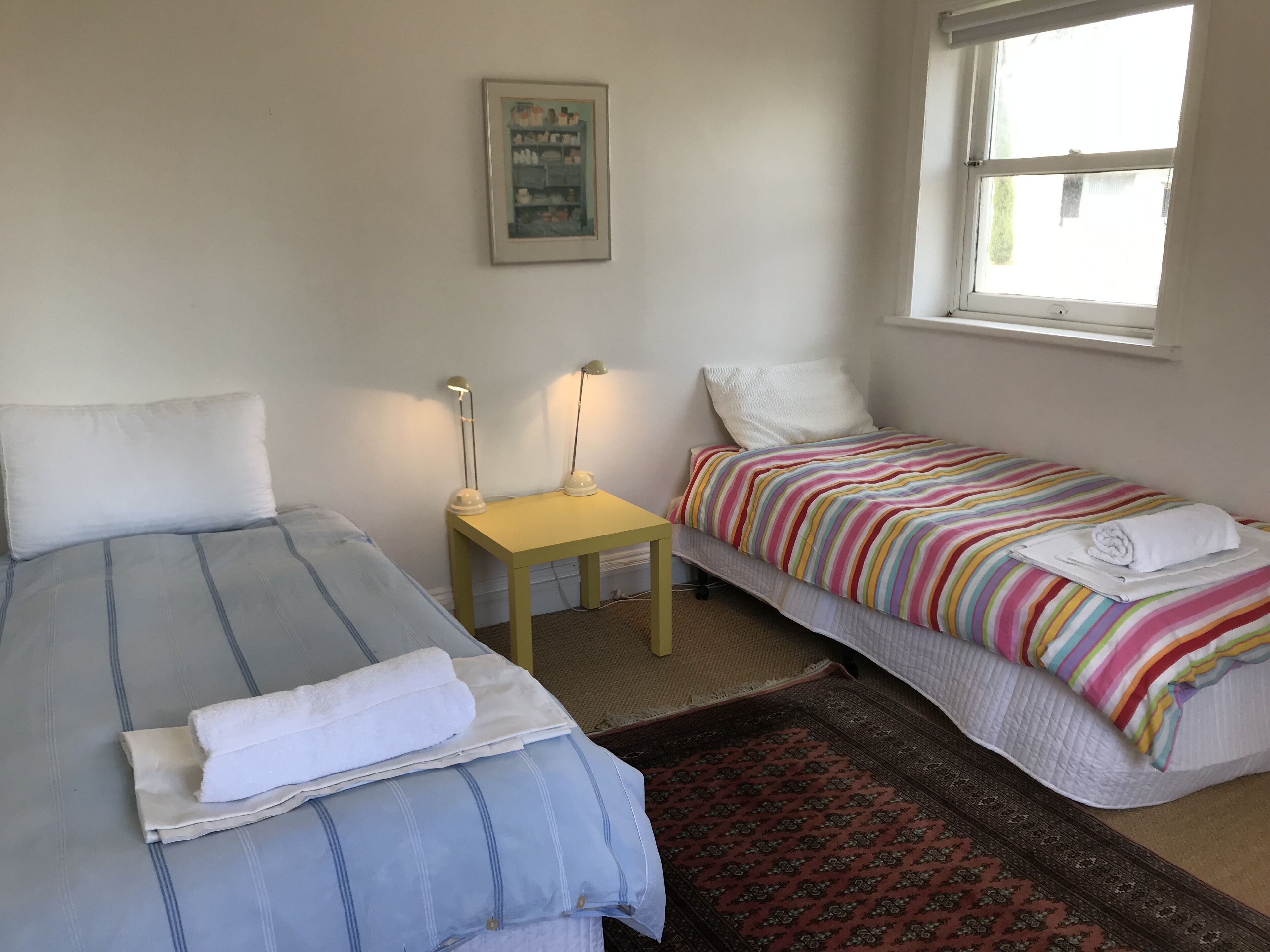 A room with two beds, a small table, a lamp, a window, and a patterned rug.