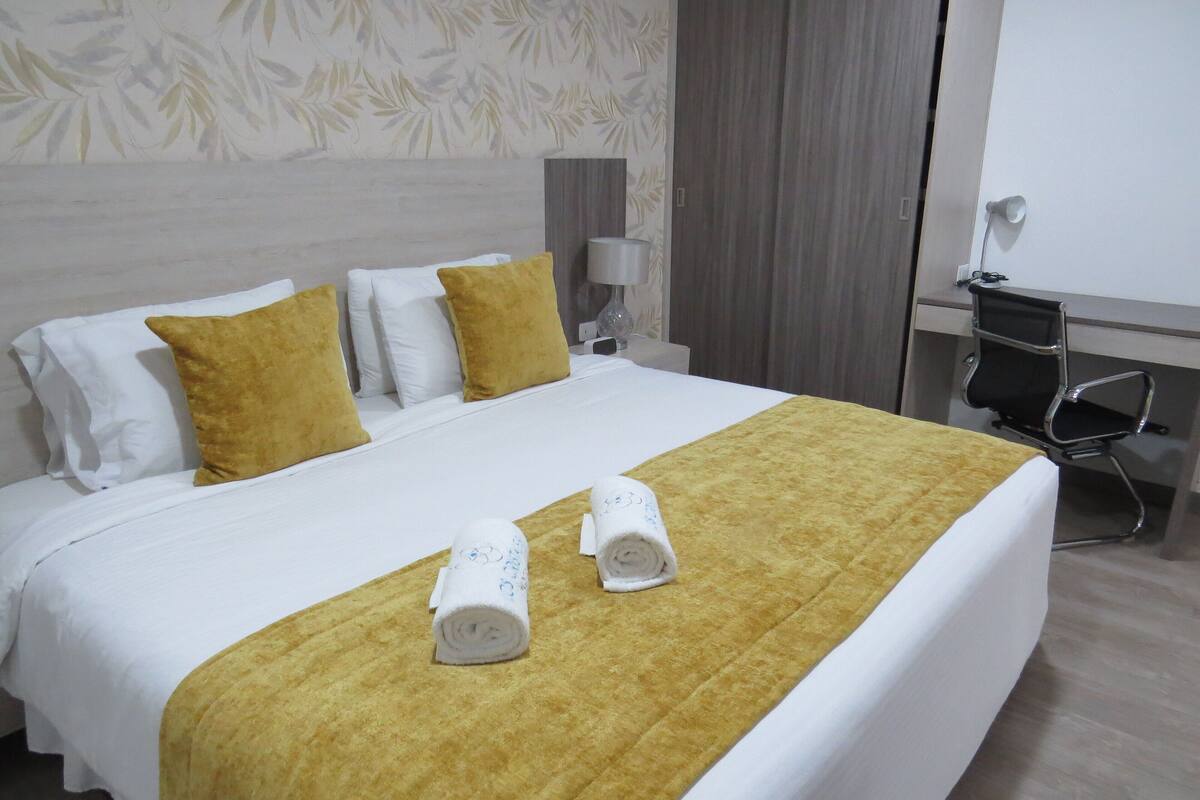 A neatly made bed with white and mustard-colored bedding, two rolled towels, and a desk with a chair and lamp.