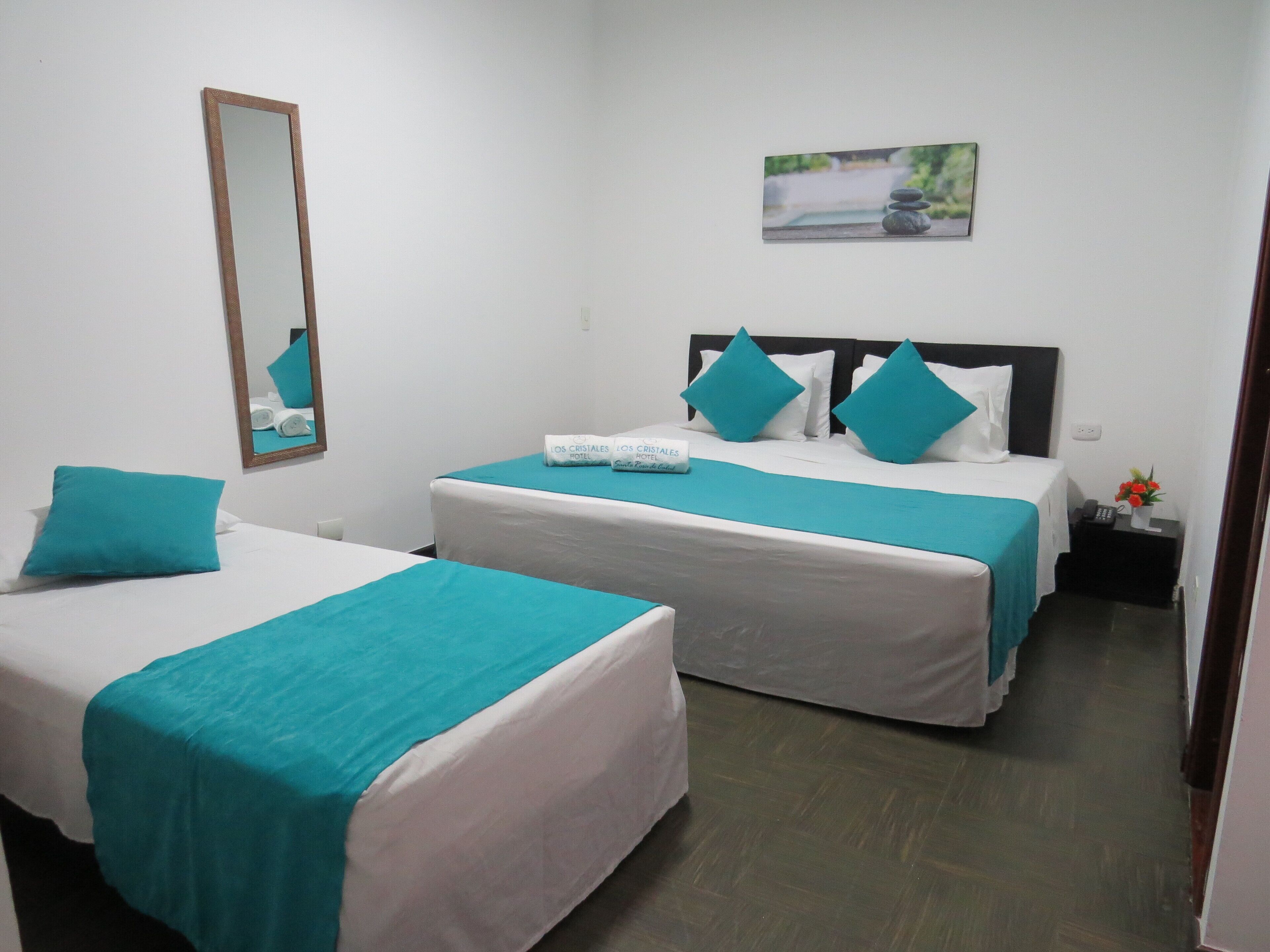 A hotel room with two beds, each with white linens and teal bedspreads. There is a small bedside table with a phone and a vase of flowers. A painting hangs on the wall above the beds.