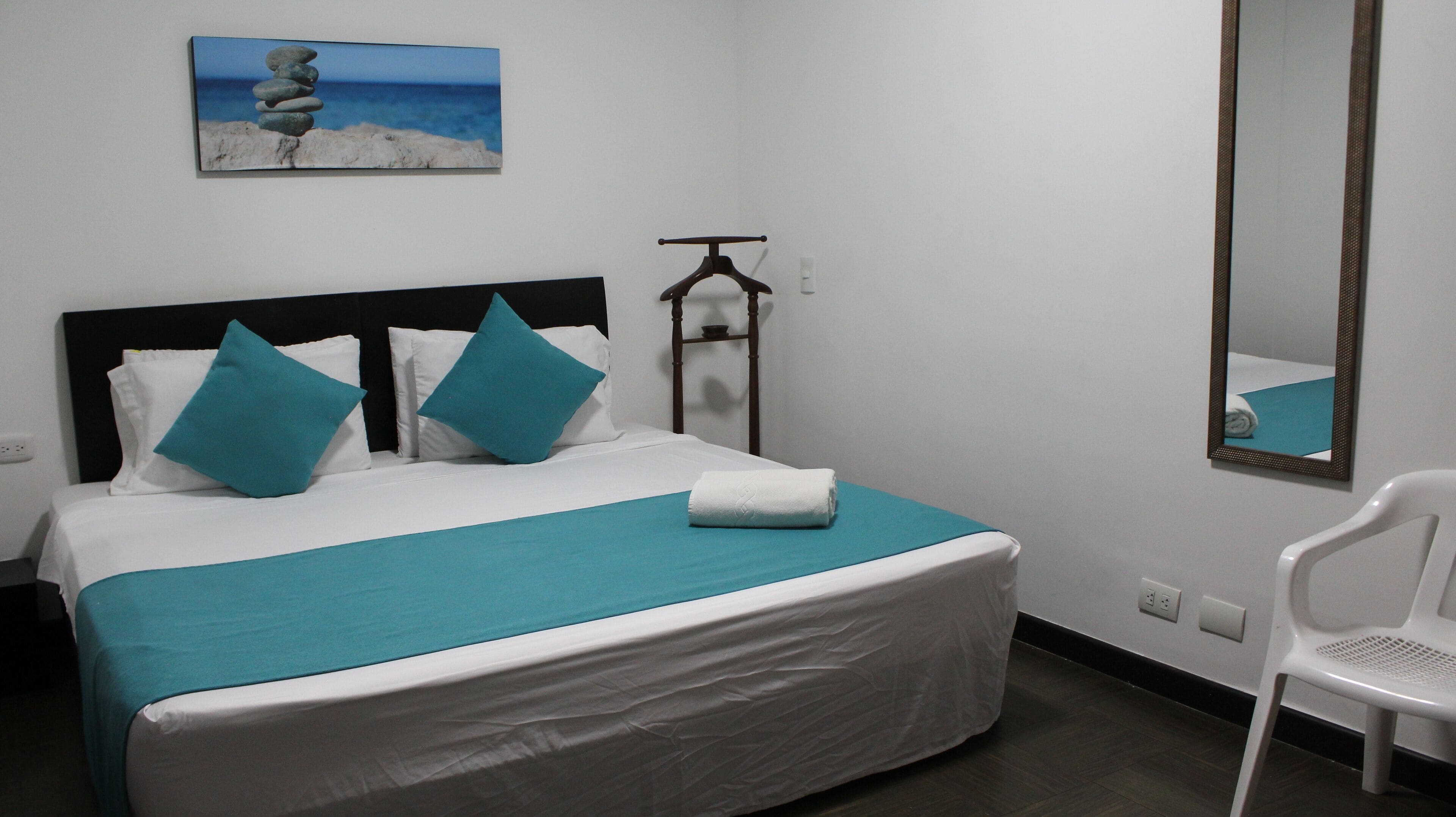 Een netjes opgemaakt bed met wit en turquoise beddengoed, een turquoise plaid en een gevouwen witte handdoek. Een witte stoel staat rechts. Een houten kapstok hangt aan de muur, en erboven hangt een schilderij van een strand.