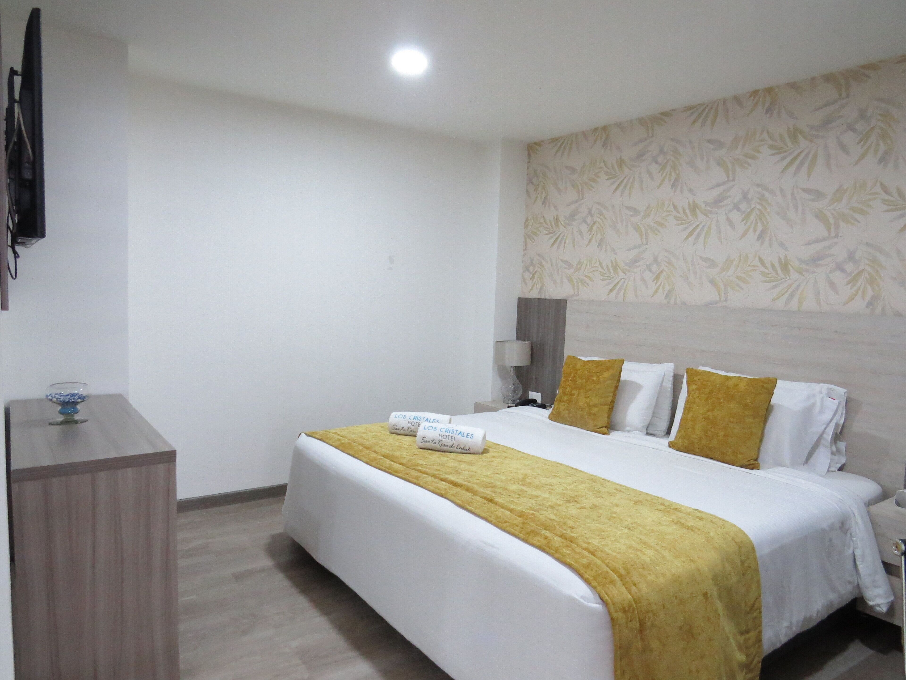 Quarto de hotel com uma cama, mesa de cabeceira, abajur e televisão fixada na parede.