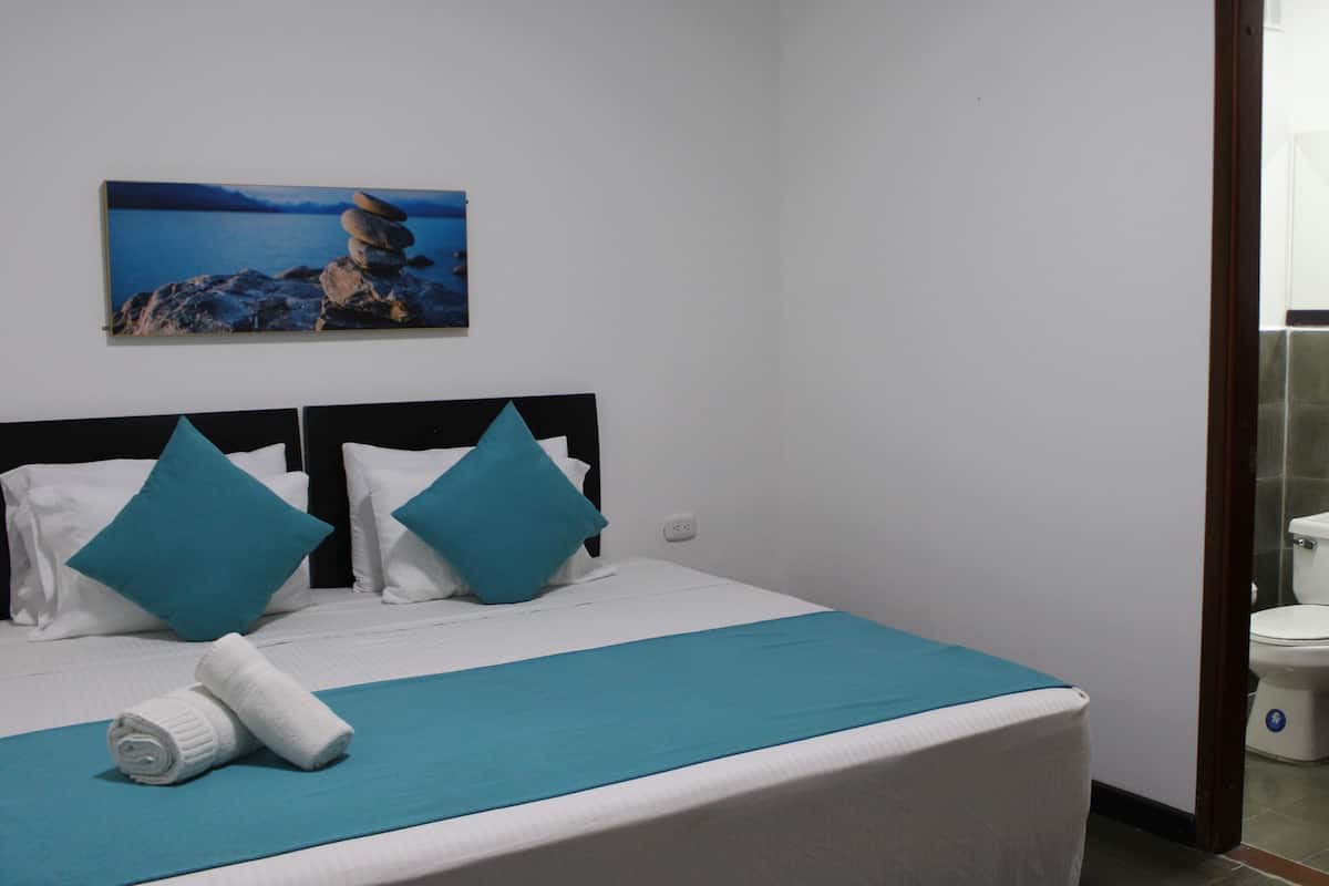 A hotel room with a bed, a painting of a coastal scene, and a view of a bathroom.