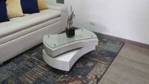 Un salon avec un canapé blanc, une table basse en verre, un tapis à motifs et un tableau abstrait encadré accroché au mur.