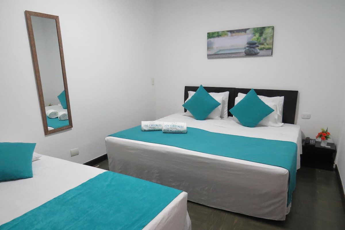 A hotel room with two beds, each with teal bedding and white pillows, a large mirror, a painting on the wall, and a small table with a phone and flowers.