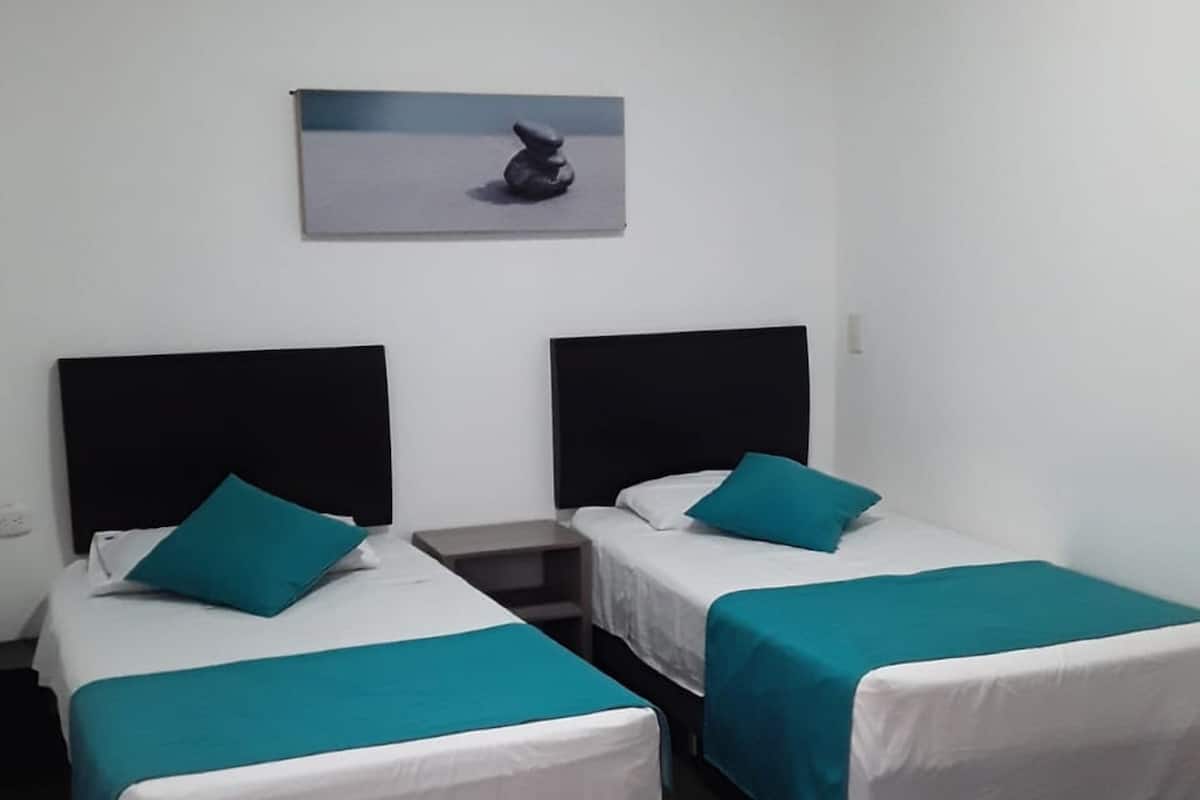 A hotel room with two single beds, each with a teal bedspread and pillow, a black headboard, and a small bedside table. There is a framed picture on the wall above the beds.