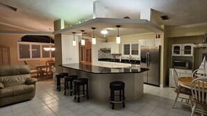 Een moderne keuken met een centraal kookeiland, barstoelen, een eethoek met een houten tafel en stoelen, en een kroonluchter.