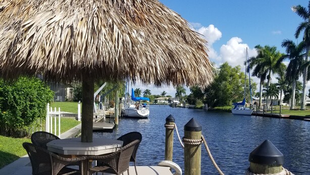 Een houten steiger met een rieten parasol, een tafel met stoelen en uitzicht op een rustig water met boten.
