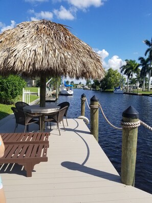 Een houten steiger met een rieten parasol, een tafel met stoelen en uitzicht op een rustig water met boten.