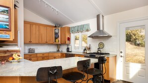 Una cocina con armarios de madera, una isla grande con taburetes de bar, una campana extractora de acero inoxidable y vistas al exterior a través de una puerta.