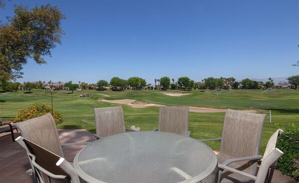 A patio with a round table and four chairs, overlooking a well-maintained golf course with sand bunkers and trees.