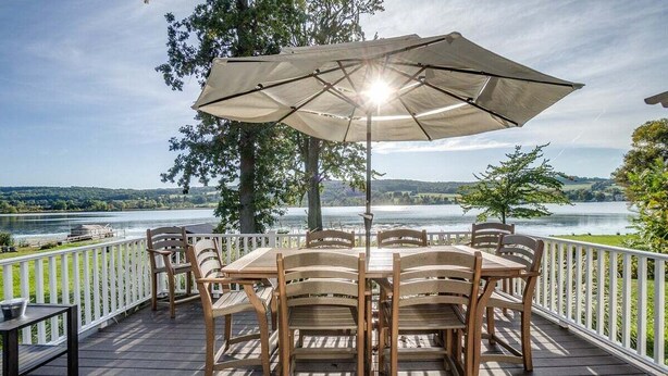 Terraza de madera con una mesa y sillas bajo un paraguas grande, con vista a un lago.
