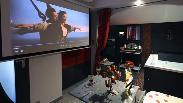 A modern hotel room with a large screen displaying a movie, a dining table set with candles and glasses, and a minibar. - Suite Rouge Passion avec jacuzzi privatif en Provence (Plan-d'Orgon)