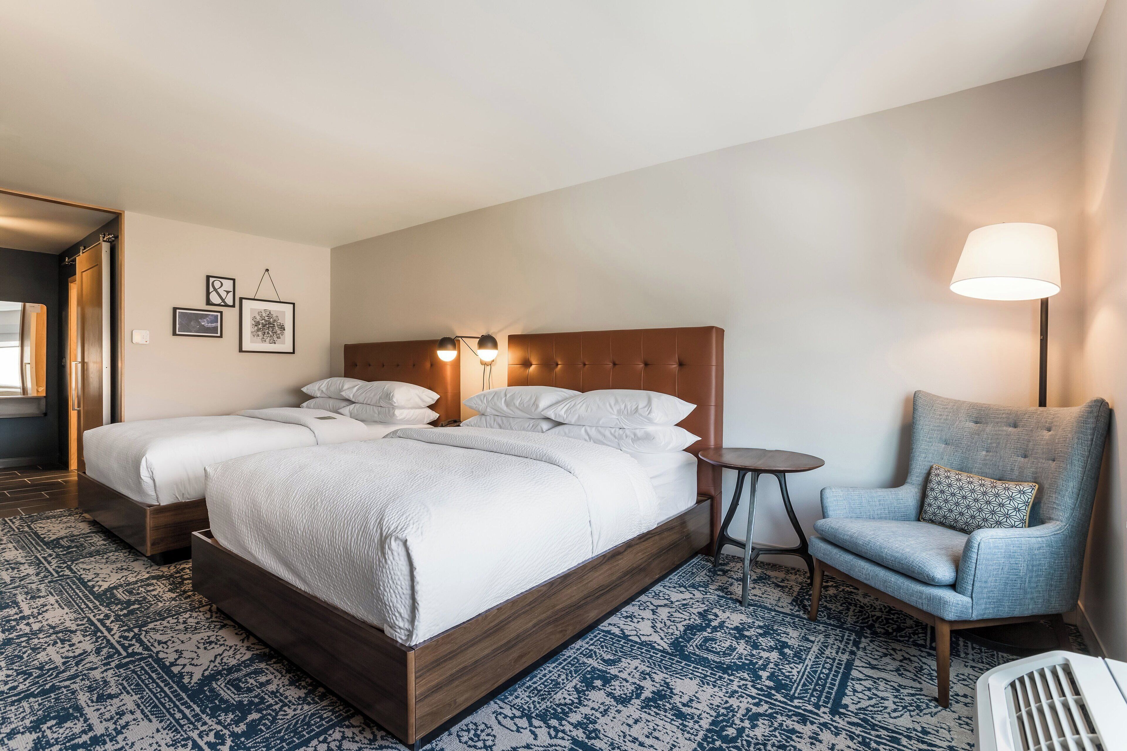 Una habitación de hotel con una cama grande, dos mesitas de noche, una silla y una lámpara.