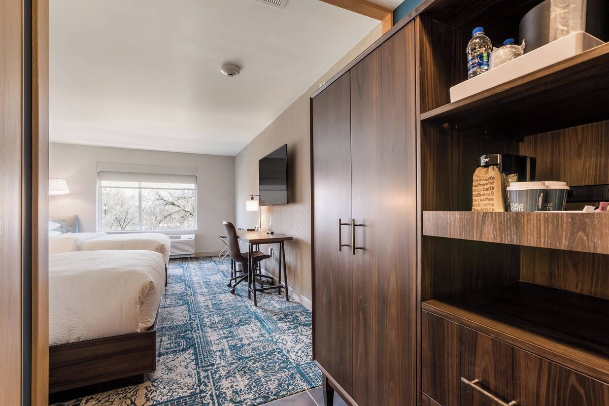 A hotel room with two beds, a desk, a chair, a TV, and a wardrobe with shelves.