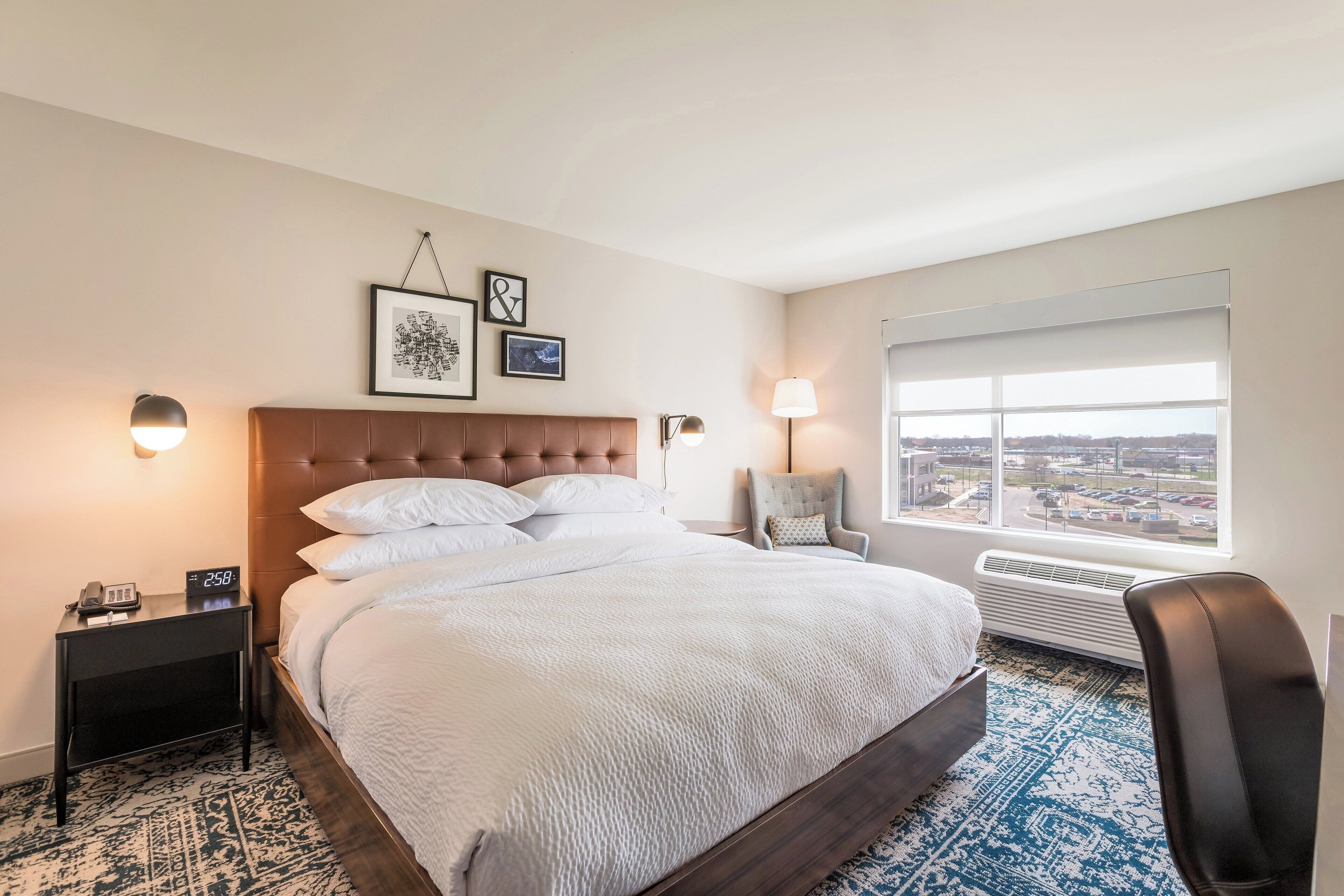 A hotel room with a large bed, a nightstand with a phone, a chair, a window with a city view, and wall-mounted artwork.