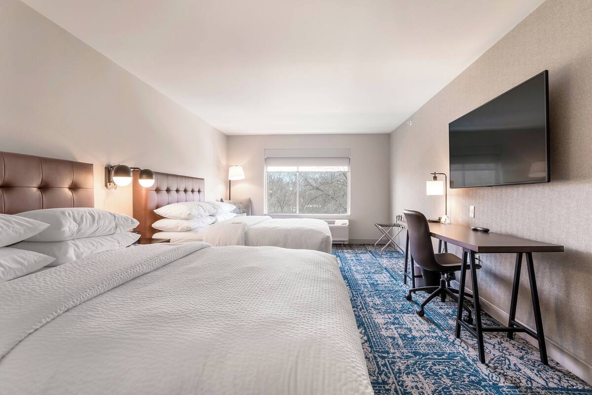 A hotel room with two beds, a desk, a chair, a flat-screen TV, and a window.