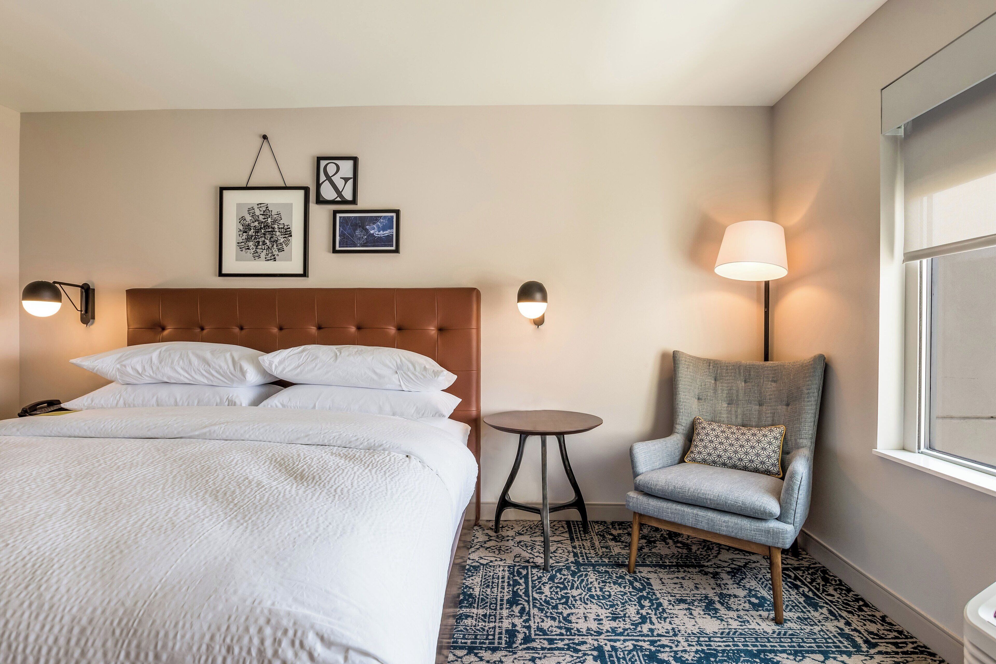 Hotellihuone, jossa on suuri sänky, pieni pyöreä pöytä, tuoli ja seinään kiinnitetyt lamput.