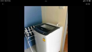 A compact, white washing machine with a control panel, situated in a small laundry area with a blue wall and a metal drying rack. - Phuket Town Budget Lodge AirconRoom1+Washer (Phuket)