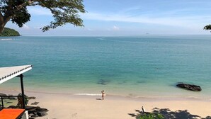 Ein Strand mit klarem türkisblauem Wasser, eine Person steht am Ufer, und ein Gebäude mit Dach und Tisch.